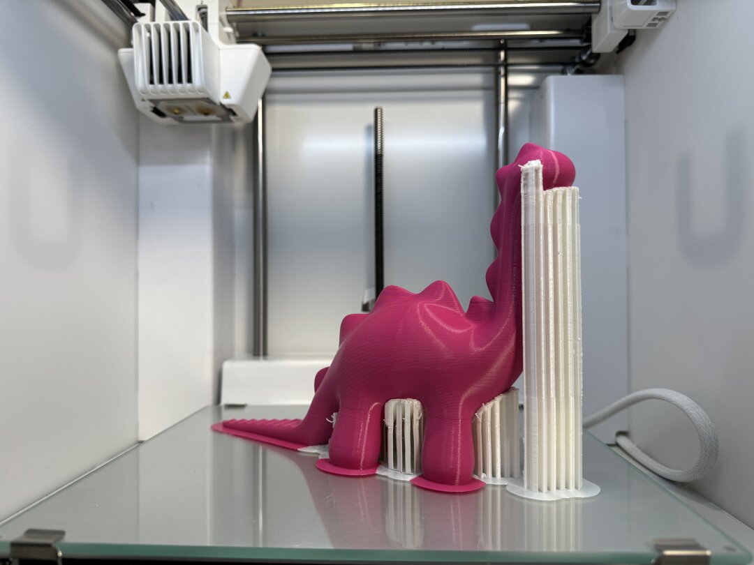 Auf dem Druckbett eines 3D-Druckers ist die Hälfte eines rosa Dinosauriers zu sehen. Weißes Stützmaterial umgibt ihn. 